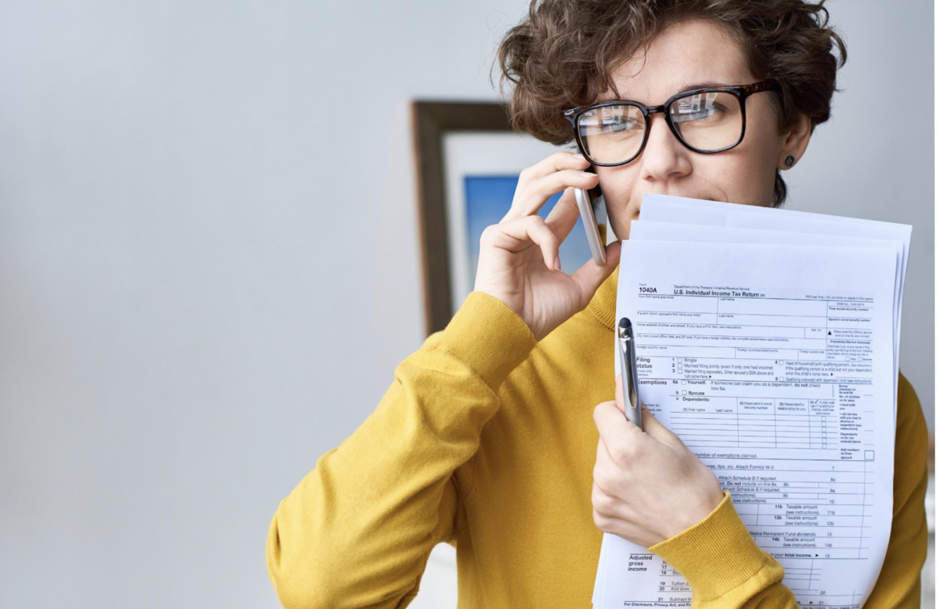 Person wearing glasses and holding tax forms while talking on the phone, symbolizing the process of seeking assistance or clarification during tax preparation.