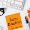 An orange sticky note with 'Taxes Deadline' written on it, next to an alarm clock, pen, and keyboard, highlighting the importance of tax deadlines.