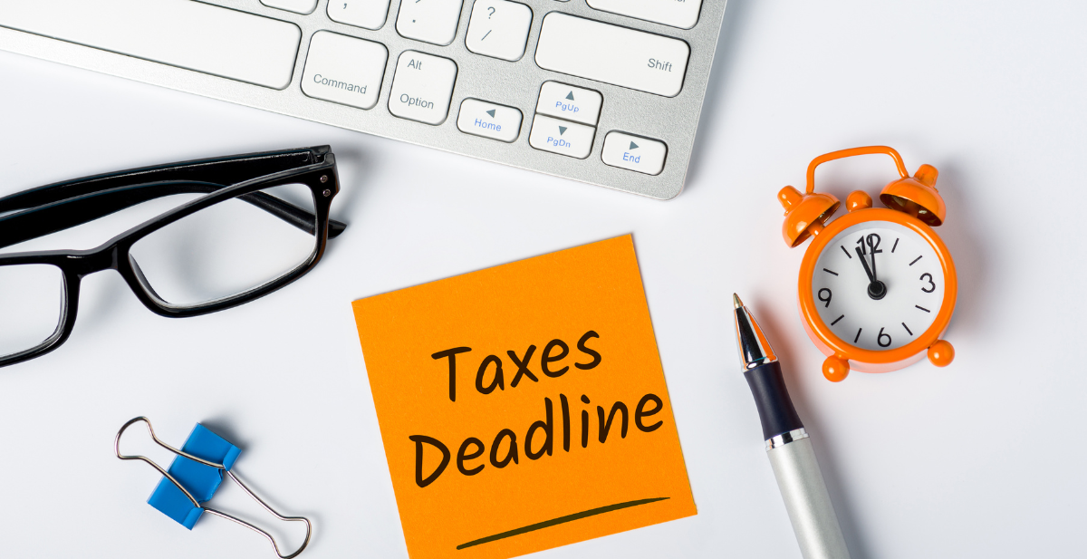 An orange sticky note with 'Taxes Deadline' written on it, next to an alarm clock, pen, and keyboard, highlighting the importance of tax deadlines.